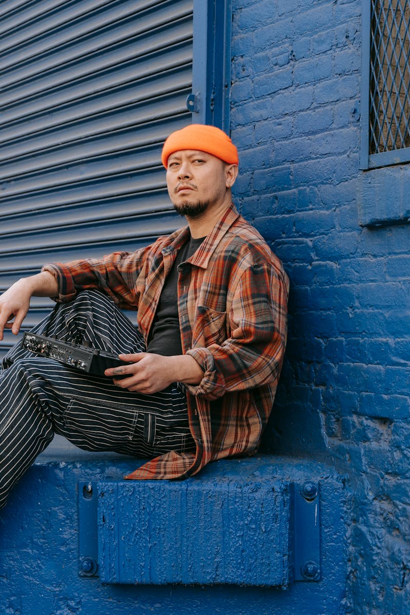 Portrait of a musician in orange beanie sitting with mixer by a blue wall, exuding urban style.